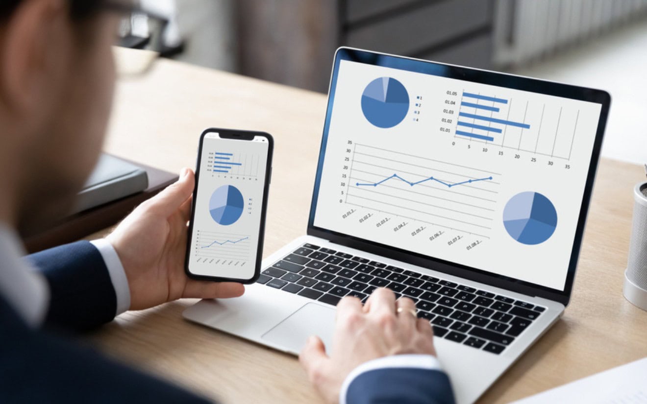 Businessman using laptop and mobile phone to view graphs on screen.